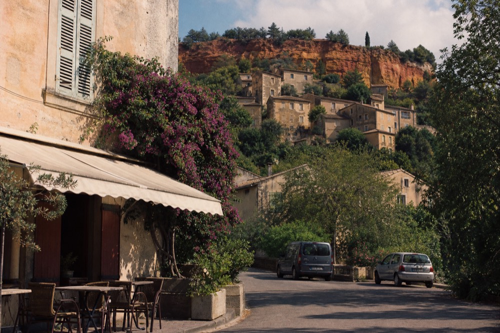 Ochre-toned village houses on a hillside with red cliffs behind, no text, no logos, no people.