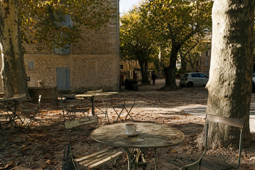 Village square with plane trees and café tables in warm afternoon light, no text, no logos, no people.