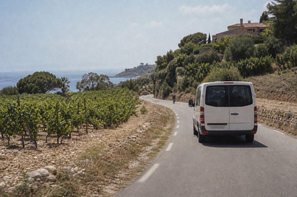 A small van on a coastal road between vineyards and the sea, no text, no logos, no people